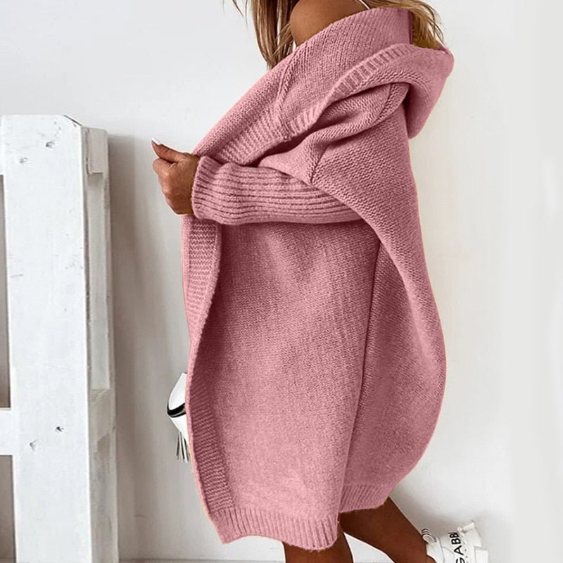 Femme portant un long cardigan rose poudré à maille épaisse avec capuche, superposé à une tenue claire, posant de profil dans un intérieur lumineux au style minimaliste.