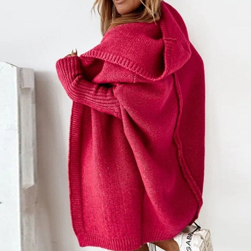 Femme portant un long cardigan rouge en maille épaisse, drapé sur les épaules et doté d'une capuche ample, dans un intérieur lumineux où l'on aperçoit un sac blanc en bas de l'image.