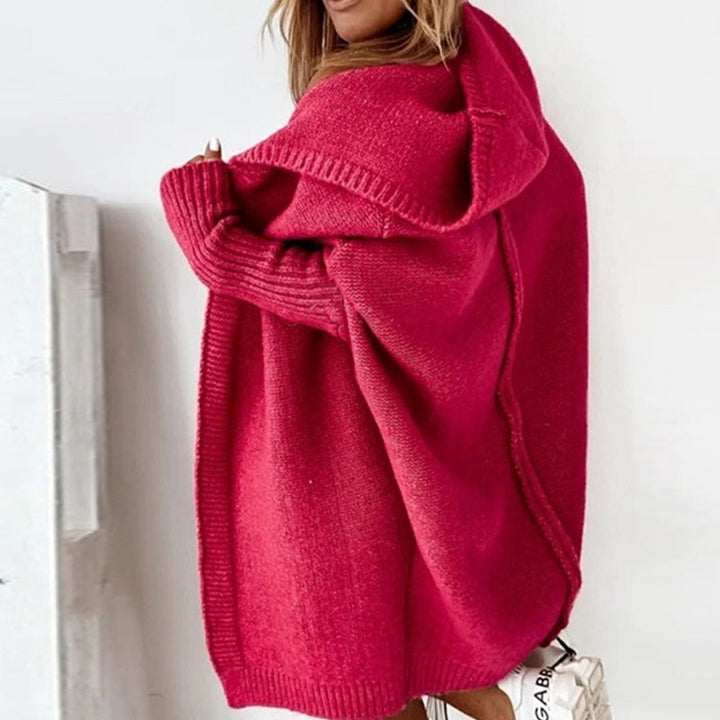 Femme portant un long cardigan rouge en maille épaisse, drapé sur les épaules et doté d'une capuche ample, dans un intérieur lumineux où l'on aperçoit un sac blanc en bas de l'image.