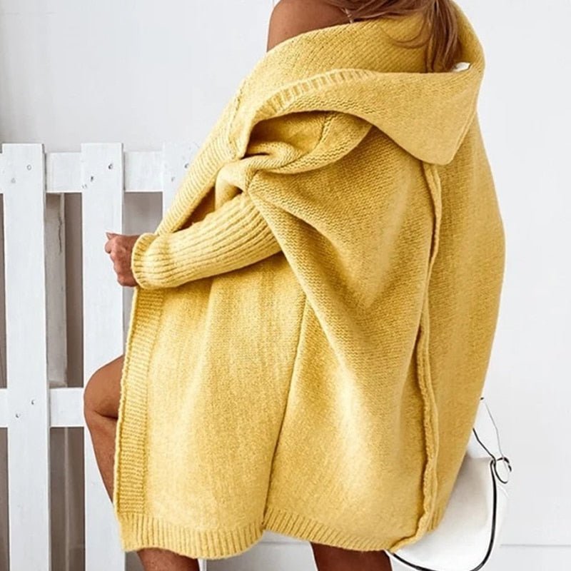Femme portant un long cardigan jaune en maille épaisse avec capuche, drapé sur les épaules, posant de profil avec un sac blanc dans un intérieur lumineux et minimaliste.
