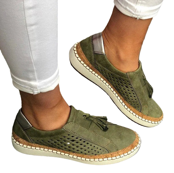 Mocassins/pantoufles plates kaki pour femme, effet suédé avec perforations latérales et pampilles, semelle beige avec liseré liège, portées avec un pantalon blanc.
