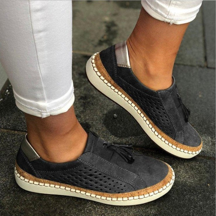 Pantoufles/mocassins plats gris anthracite effet suédé, avec perforations latérales et pampilles, semelle beige avec liseré liège, portées en extérieur avec un pantalon blanc.