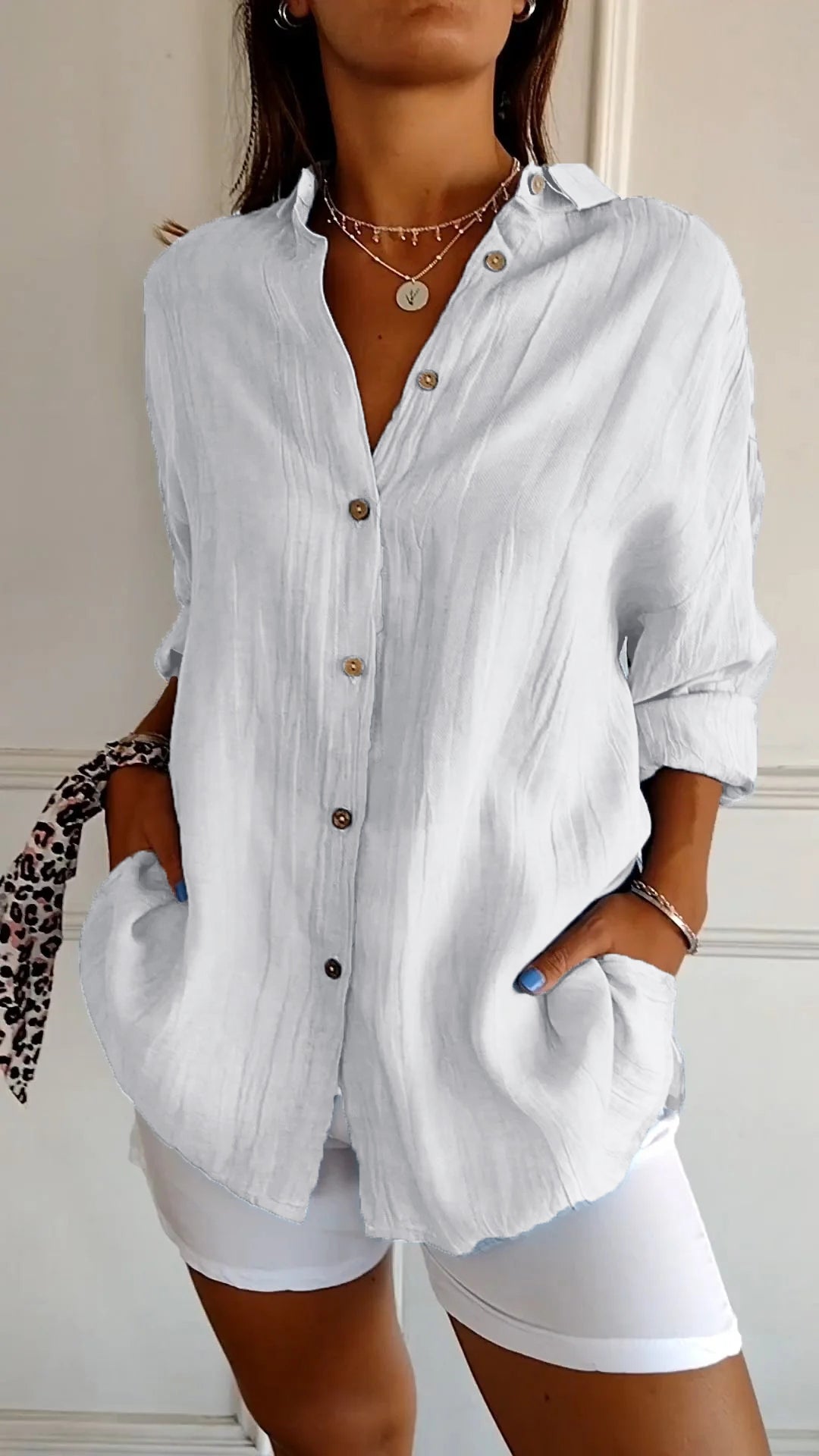 Chemise plissée pour femme blanche, coupe ample avec col ouvert et manches retroussées, boutonnage devant, portée avec un short blanc.