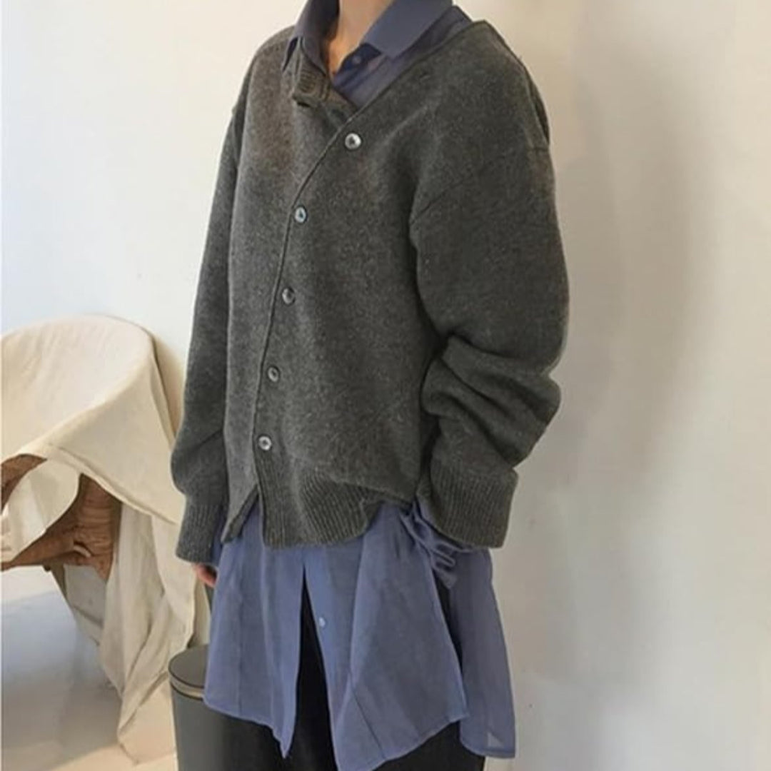 Femme portant un cardigan gris en maille, avec des boutons gris et un col en V. Elle porte une chemise bleue sous le cardigan, créant un look superposé moderne et décontracté. Elle pose de profil contre un mur blanc, affichant un style élégant et confortable.