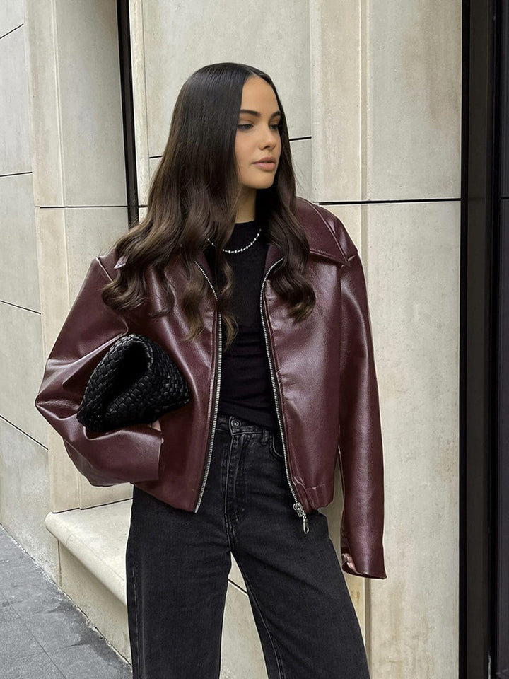 Femme portant une veste en simili cuir bordeaux avec col large et fermeture zippée, associée à un haut noir, un jean noir et un sac à main noir, pour un look moderne et élégant.