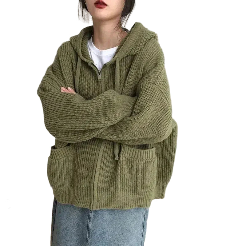 Une femme porte un cardigan vert en grosse maille zippée, à capuche et grandes poches avant. Sa coupe ample lui confère un style décontracté et confortable. Il est associé à un t-shirt blanc et une jupe en jean. Parfait pour un look moderne et cosy au quotidien.