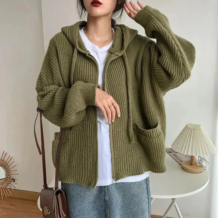 Une femme porte un cardigan vert en grosse maille, à capuche et grandes poches avant, ouvert sur un t-shirt blanc. Ce vêtement ample offre un style décontracté et chaud, idéal pour un look d'automne moderne et confortable.