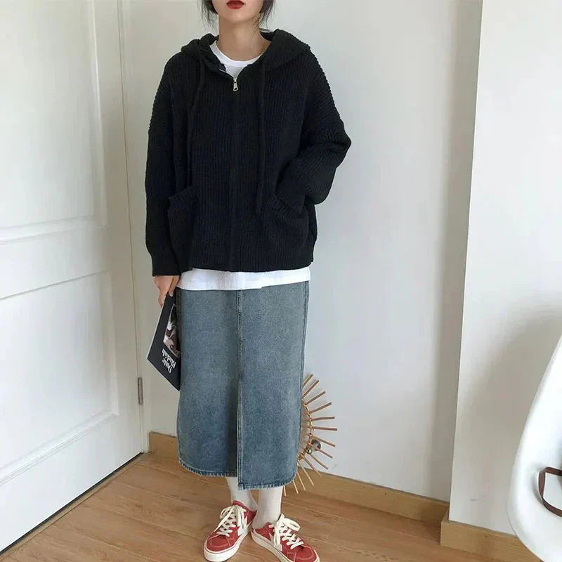 Femme habillée d’un cardigan noir en maille épaisse avec capuche et poches avant, porté sur un t-shirt blanc et une jupe midi en jean. La tenue est complétée par des baskets rouges et blanches, créant un look décontracté et moderne parfait pour la mi-saison.