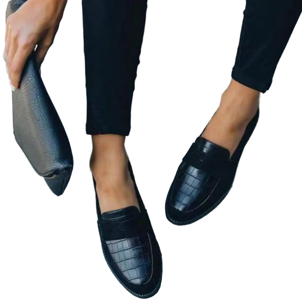 Chaussures d’été pour femme de type mocassins, en cuir noir à texture effet croco, avec bout arrondi et coupe plate, portées avec un pantalon noir pour un style élégant et moderne.