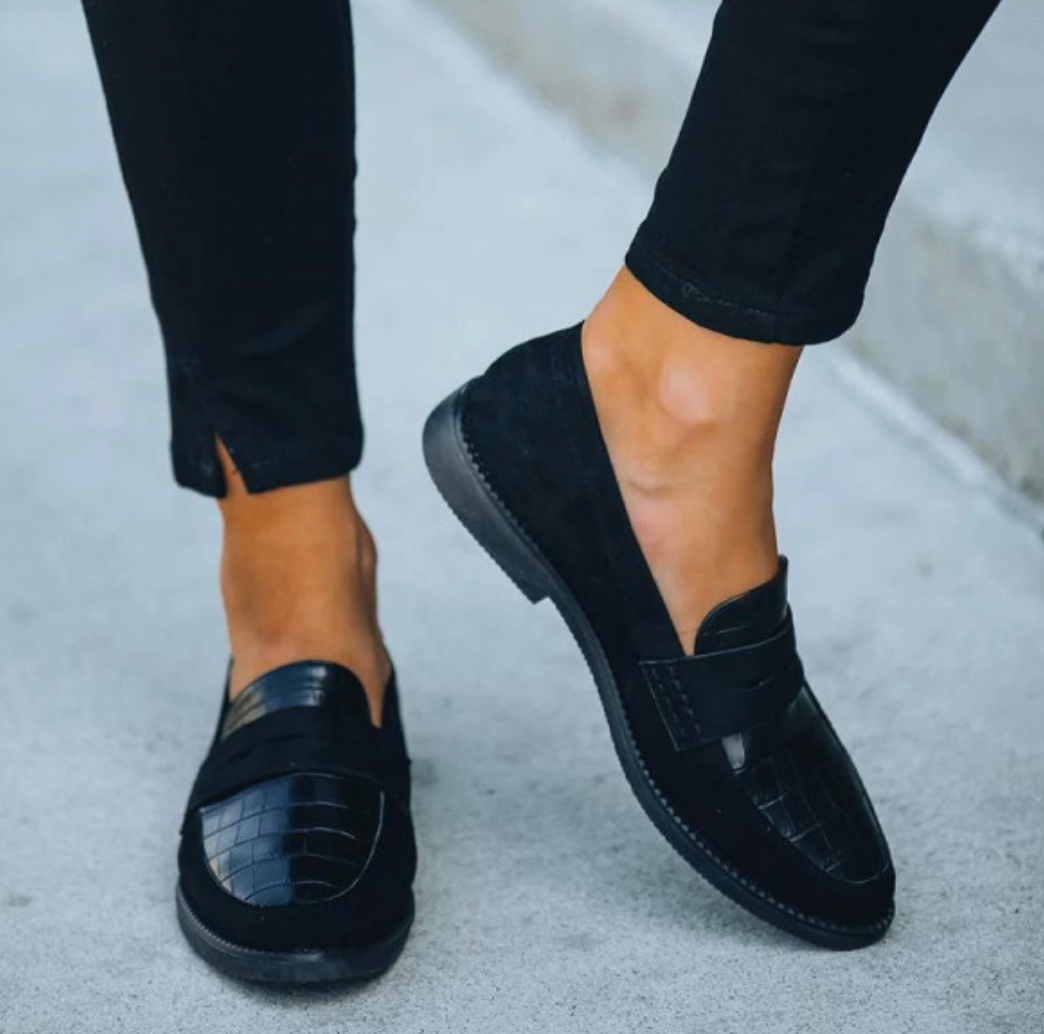 Mocassins d’été pour femme en cuir noir, avec empiècement texturé effet croco et semelle plate, portés en mouvement avec un pantalon noir pour un look moderne et confortable.