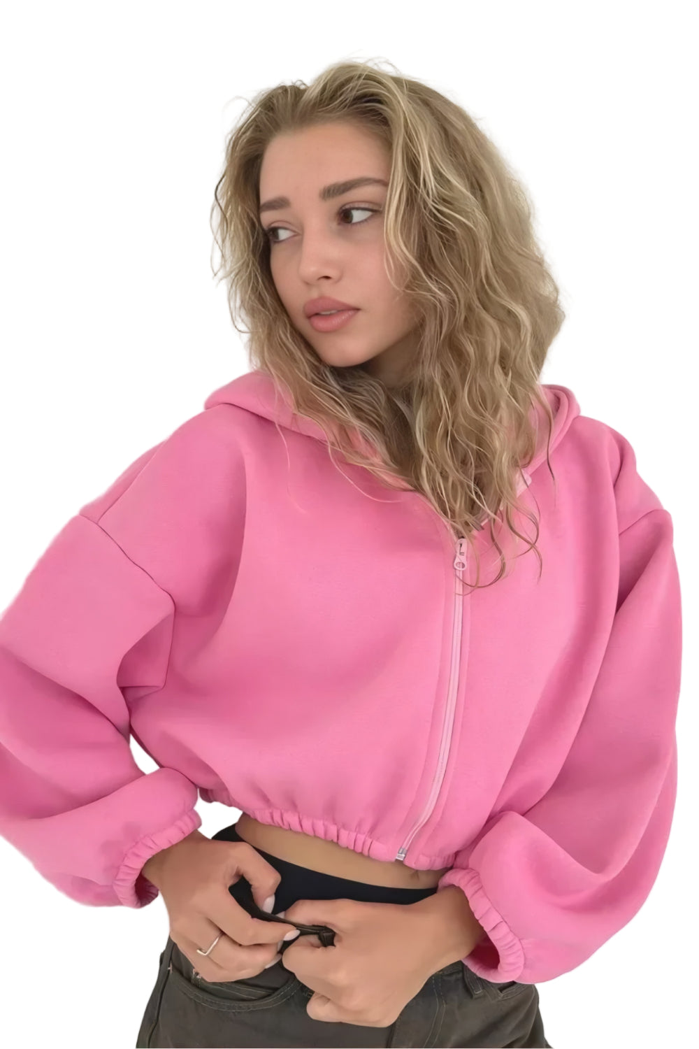 Une femme vêtue d'un sweat-shirt court rose à manches bouffantes ajuste sa ceinture en posant légèrement de profil. Son allure est douce et naturelle sur un fond neutre et épuré.