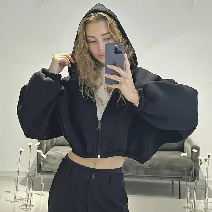 Jeune femme portant un sweat à capuche noir court, avec des manches volumineuses et une coupe moderne, posant devant un miroir. Elle porte un pantalon noir et un haut blanc, et prend un selfie en ajustant la capuche.