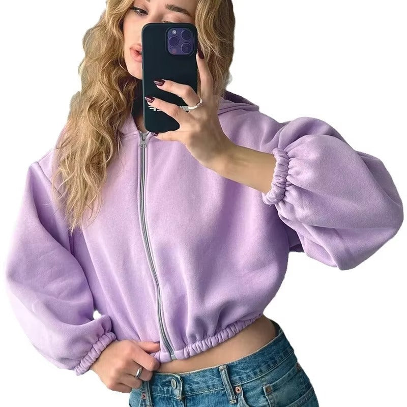 Une femme vêtue d'un sweat-shirt court violet à manches bouffantes et à la coupe moderne prend un selfie. Elle porte un jean bleu et affiche un style décontracté mais tendance, accessoirisé de détails simples comme une bague et un vernis à ongles foncé.
