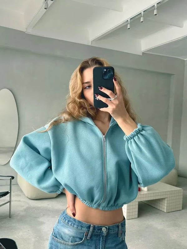 Une femme vêtue d'un sweat-shirt court bleu à manches bouffantes et à la coupe moderne prend un selfie devant un miroir. Elle porte un jean bleu et affiche un look à la fois décontracté et tendance, avec une pose naturelle et un fond minimaliste.