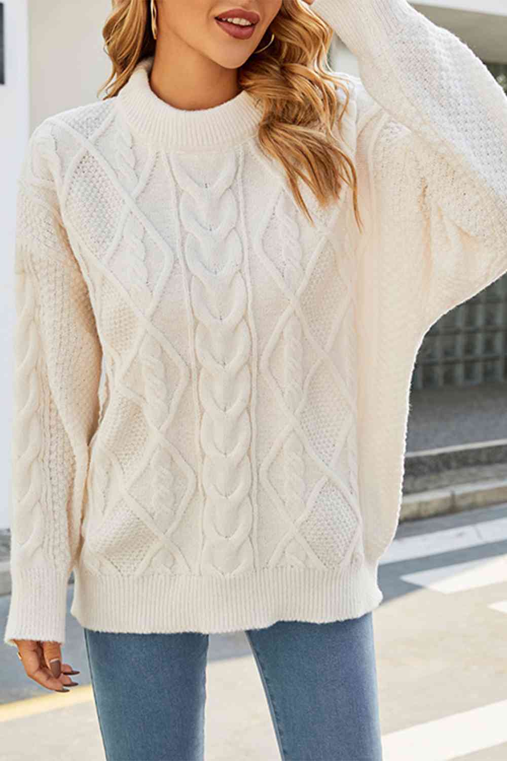 Femme portant un pull en maille torsadée blanc cassé à col montant, au style ample et décontracté. Le tricot présente un motif en losanges et torsades sur le devant et les manches, créant une texture élégante et moderne. Le pull est associé à un jean bleu clair, offrant un look confortable et chic, parfait pour la saison automne-hiver. Les cheveux ondulés et les accessoires dorés complètent une tenue féminine et chaleureuse sous une lumière naturelle.