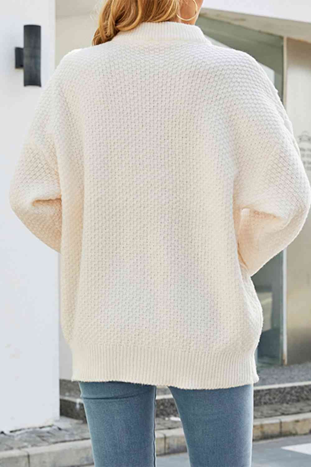 Vue de dos d'une femme portant un pull blanc à col montant en maille, dans une coupe ample et décontractée. Le dos du pull présente une maille épaisse et régulière, soulignant la douceur et la qualité du tissu. Il est associé à un jean bleu clair pour un look moderne et confortable. La scène se déroule en extérieur, sous une lumière naturelle qui accentue la teinte douce et élégante du vêtement.
