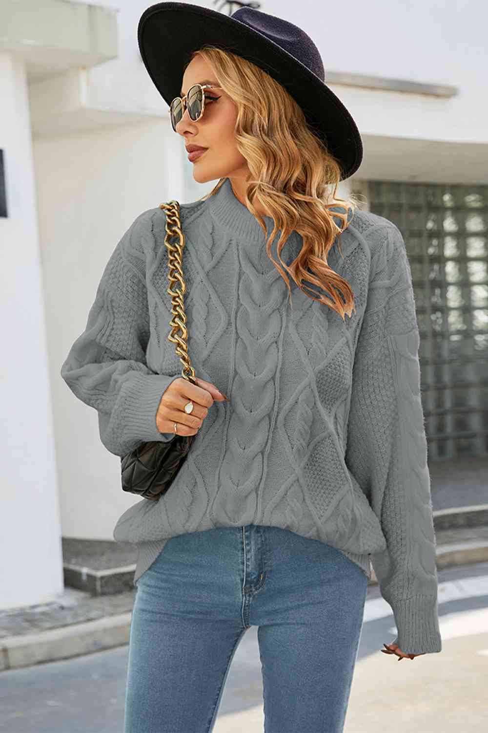Femme élégante portant un pull en maille torsadée gris à col montant, au style ample et moderne. Le tricot présente des motifs en losanges et torsades sur le devant et les manches, apportant texture et raffinement. Elle associe le pull à un jean bleu clair, un sac à chaîne dorée, des lunettes de soleil rondes et un chapeau noir à larges bords. La tenue, chic et décontractée, est mise en valeur par une lumière naturelle dans un cadre urbain minimaliste.