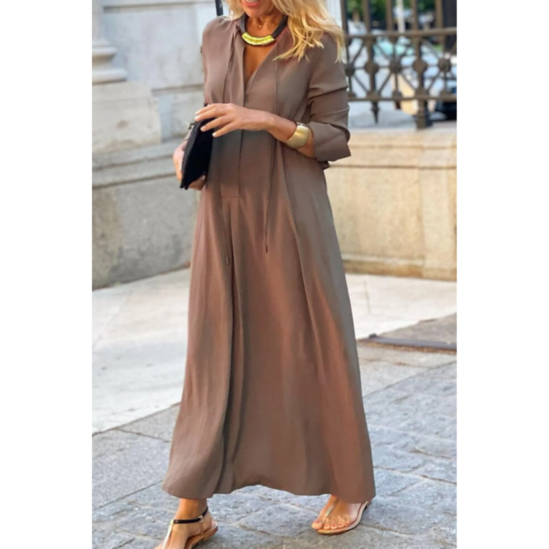 Femme portant la robe maxi d’été Nadège, longue robe marron taupe fluide à manches 3/4 et col en V, coupe ample et élégante, portée avec des sandales et des bijoux dorés  style moderne et chic en extérieur.