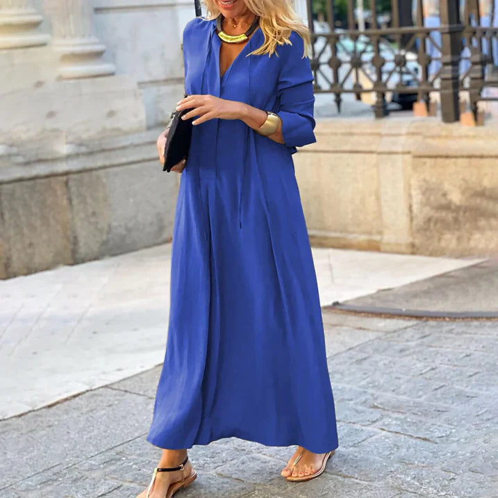 Femme portant la robe maxi d’été Nadège, longue robe bleue fluide à manches 3/4 et col en V, coupe ample et élégante, portée avec des sandales et des accessoires dorés  look moderne et chic en extérieur.