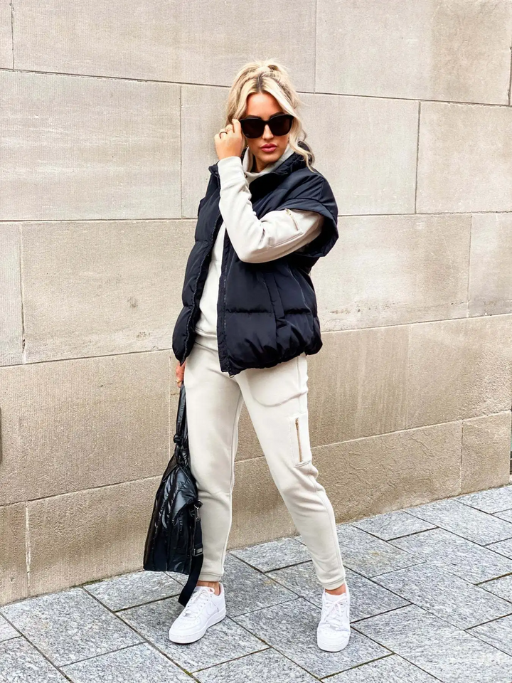 Femme moderne portant un gilet matelassé noir sans manches sur un ensemble beige clair composé d’un sweat et d’un pantalon de jogging. Elle accessoirise sa tenue avec de grandes lunettes de soleil noires, un sac à main noir brillant et des baskets blanches. La scène se déroule en extérieur devant un mur de pierre, créant un look urbain, confortable et tendance pour la saison hivernale.