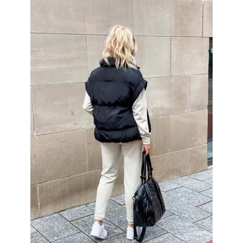 Femme vue de dos portant un gilet matelassé noir sans manches sur un ensemble beige clair composé d’un sweat et d’un pantalon de jogging. Elle tient un grand sac noir à la main et porte des baskets blanches. Ses cheveux blonds tombent en ondulations sur ses épaules. Le style est décontracté, moderne et parfait pour une promenade hivernale en ville.