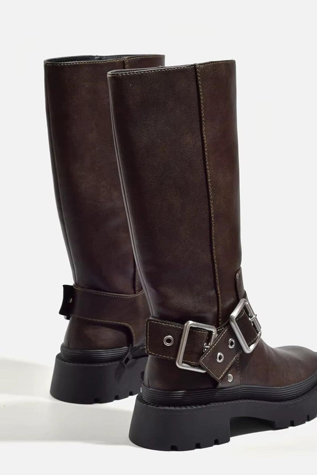 Bottes hautes pour femme en cuir synthétique marron, vues de dos et de profil, avec semelles crantées noires épaisses. Elles sont dotées d'une boucle en métal argenté à la cheville et de surpiqûres contrastantes qui soulignent leur finition soignée. Le cuir légèrement satiné et leur forme robuste leur confèrent un style moderne et tendance. Le fond clair met en valeur les détails du design et la solidité de la semelle.