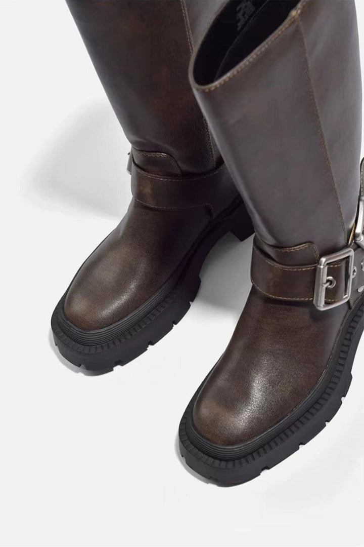 Gros plan sur une paire de bottes hautes pour femme en cuir synthétique marron, mettant en valeur la texture lisse et la finition soignée de la matière. Le modèle présente une large semelle crantée noire et une boucle métallique argentée sur le côté, lui conférant une allure à la fois moderne et robuste. Le cuir légèrement brillant souligne la qualité du produit, tandis que le fond clair met en valeur les détails du bout arrondi et du talon épais.