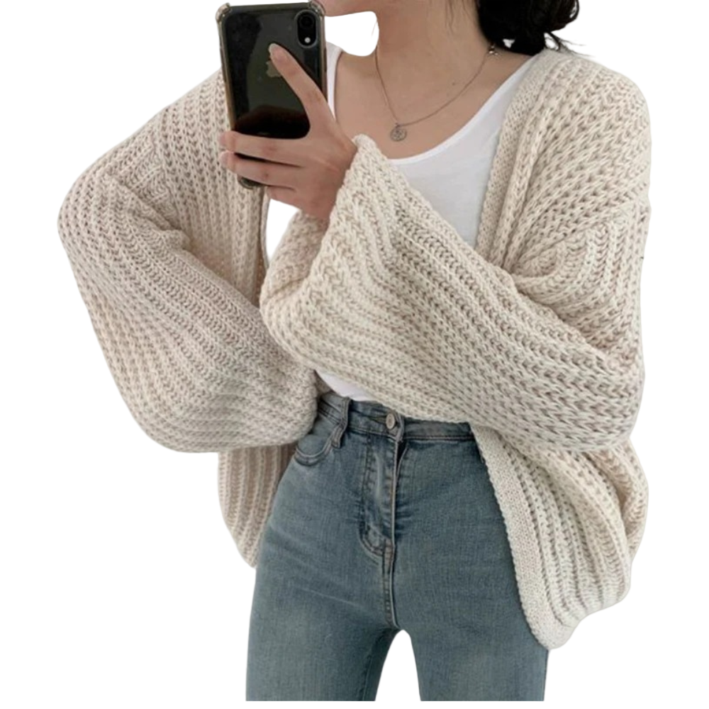 Une femme portant un cardigan blanc oversize en maille à manches larges, associé à un débardeur blanc et un jean taille haute, tient un smartphone pour se photographier devant un fond neutre.