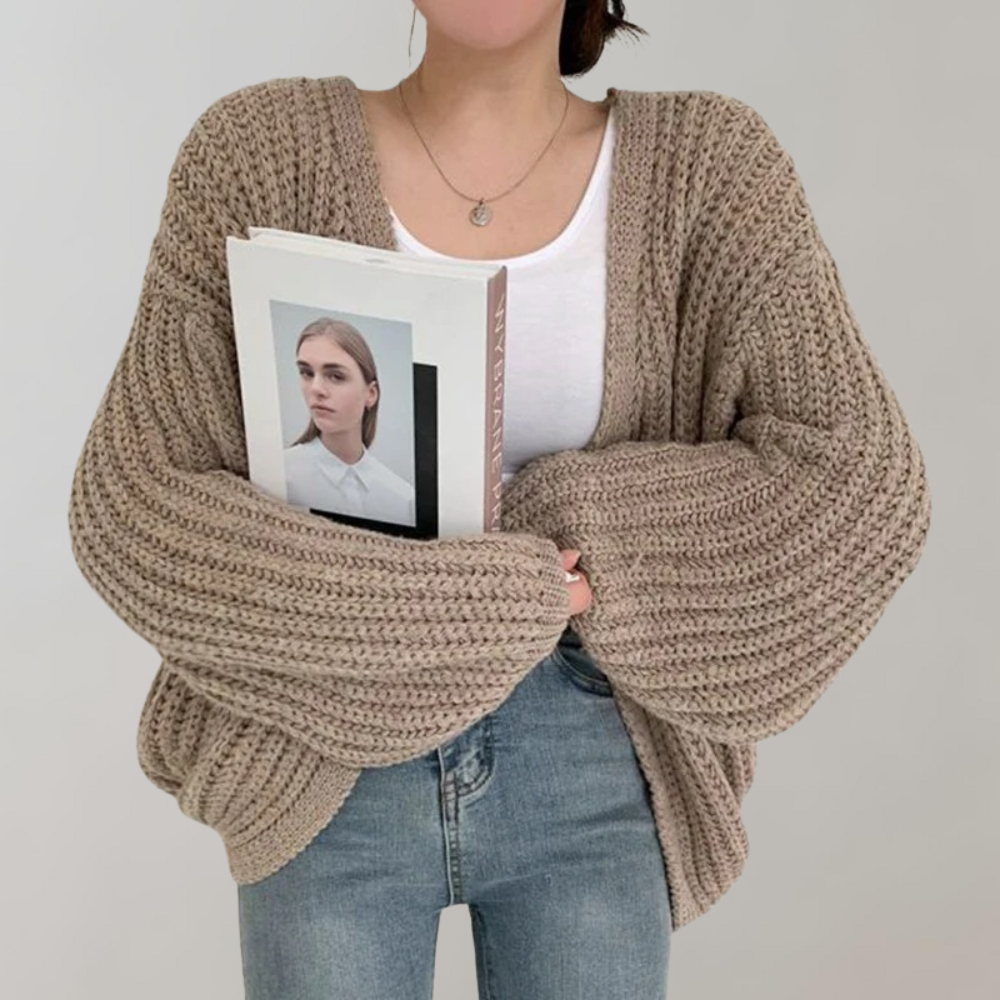 Une femme portant un cardigan kaki oversize en maille à manches larges, associé à un débardeur blanc et un jean taille haute, tient un livre contre elle et pose devant un fond neutre.