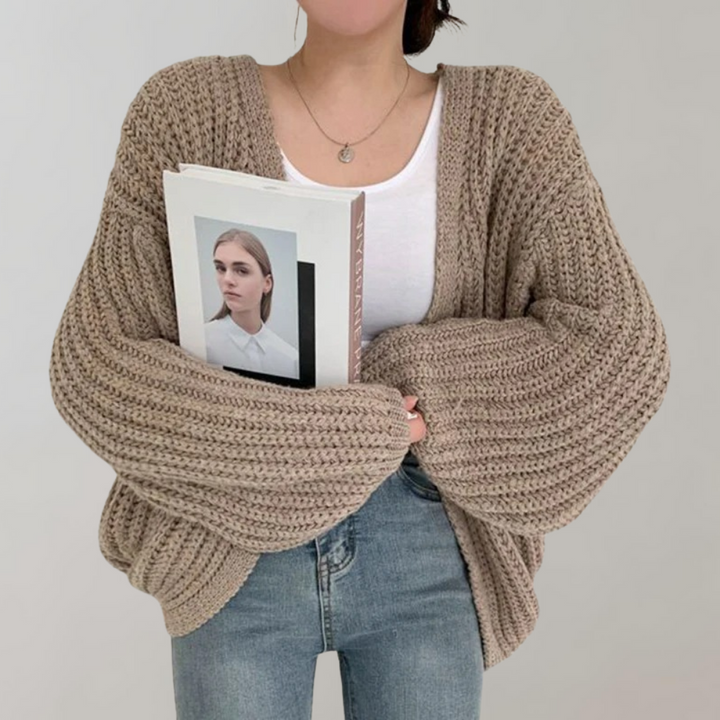Une femme portant un cardigan kaki oversize en maille à manches larges, associé à un débardeur blanc et un jean taille haute, tient un livre contre elle et pose devant un fond neutre.