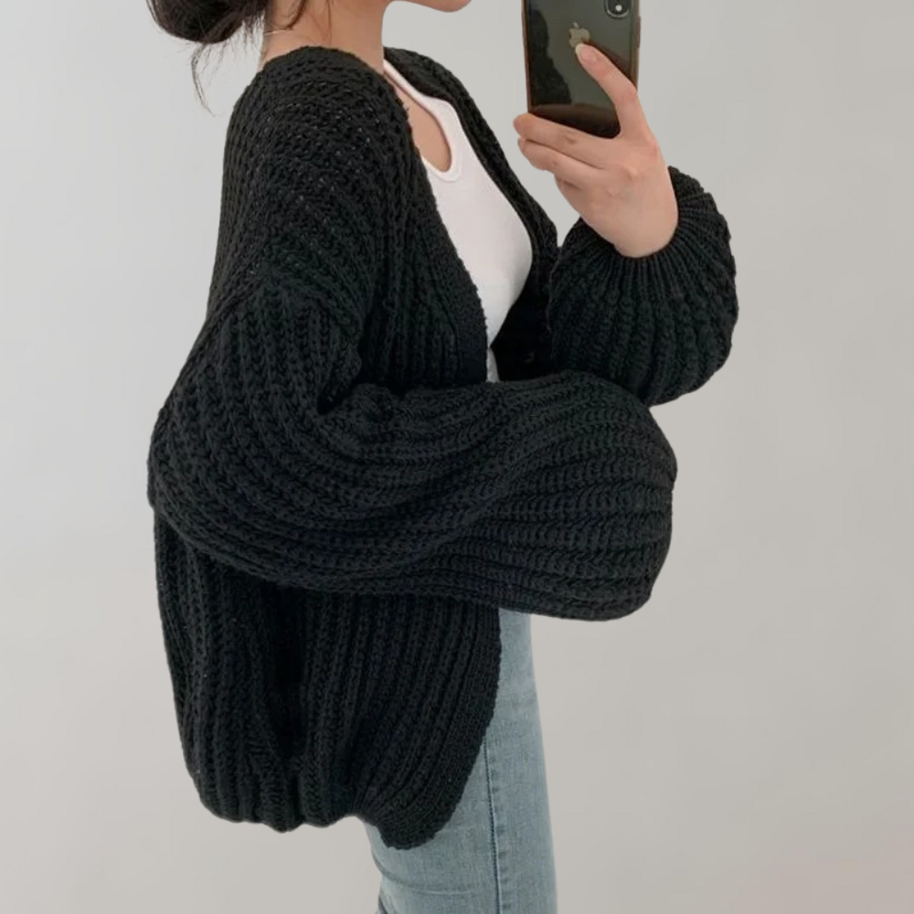 Femme portant un cardigan oversize en maille noire, aux manches très larges et texturées, associé à un débardeur blanc et un jean, prenant une photo en miroir devant un fond neutre.