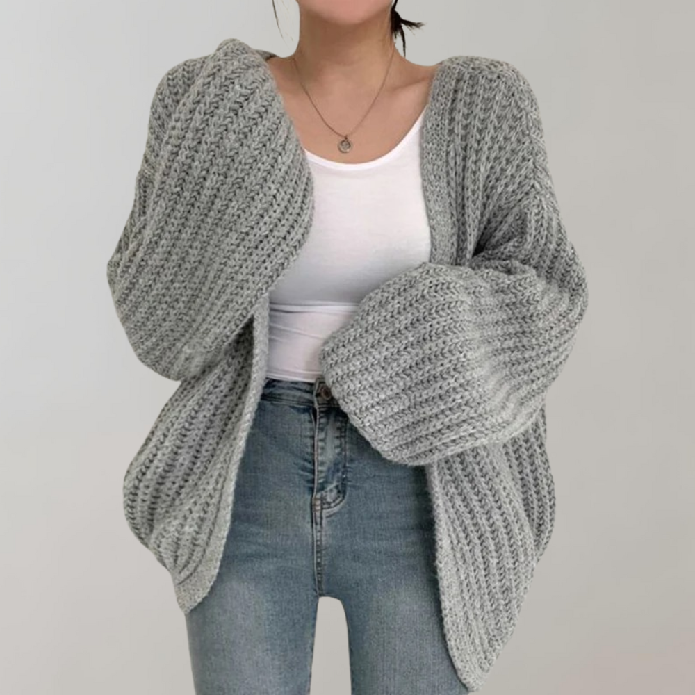 Femme portant un cardigan oversize en maille grise, aux manches volumineuses et texturées, associé à un débardeur blanc et un jean taille haute, posant devant un fond neutre.