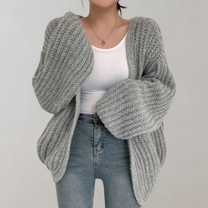 Femme portant un cardigan oversize en maille grise, aux manches volumineuses et texturées, associé à un débardeur blanc et un jean taille haute, posant devant un fond neutre.