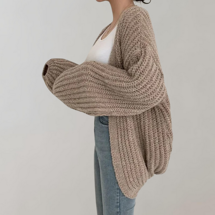 Femme de profil portant un cardigan kaki oversize en maille à manches longues et amples, associé à un débardeur blanc et un jean taille haute, posant devant un fond neutre.
