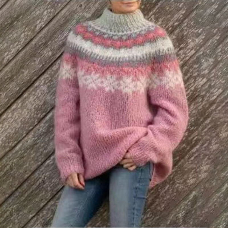 Femme portant un pull d’hiver en maille épaisse rose avec un col montant et un motif jacquard aux nuances de gris, blanc et rose, posant devant une paroi en bois pour un look chaud et confortable.