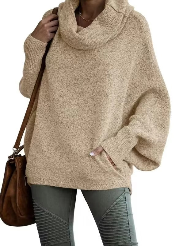 Une femme porte un pull beige oversize à manches chauve-souris et à large col drapé. La maille, douce et épaisse, est idéale pour l'hiver. Elle l'associe à un pantalon kaki ajusté et à un sac en cuir marron pour un look hivernal à la fois décontracté et élégant.
