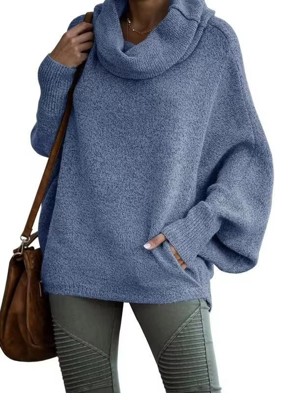 Une femme porte un pull bleu ample à manches chauve-souris et à large col drapé. Le tissu est doux et confortable, idéal pour l'hiver. Elle l'associe à un pantalon kaki ajusté et à un sac en cuir marron pour une allure à la fois décontractée et élégante.
