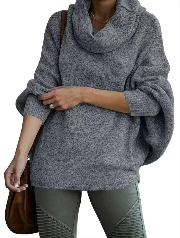Une femme porte un pull gris foncé à manches chauve-souris amples et à grand col drapé. Le tissu doux et fluide lui confère un style confortable et moderne. Elle complète sa tenue avec un pantalon kaki texturé et un sac en cuir marron, parfait pour un look hivernal décontracté.