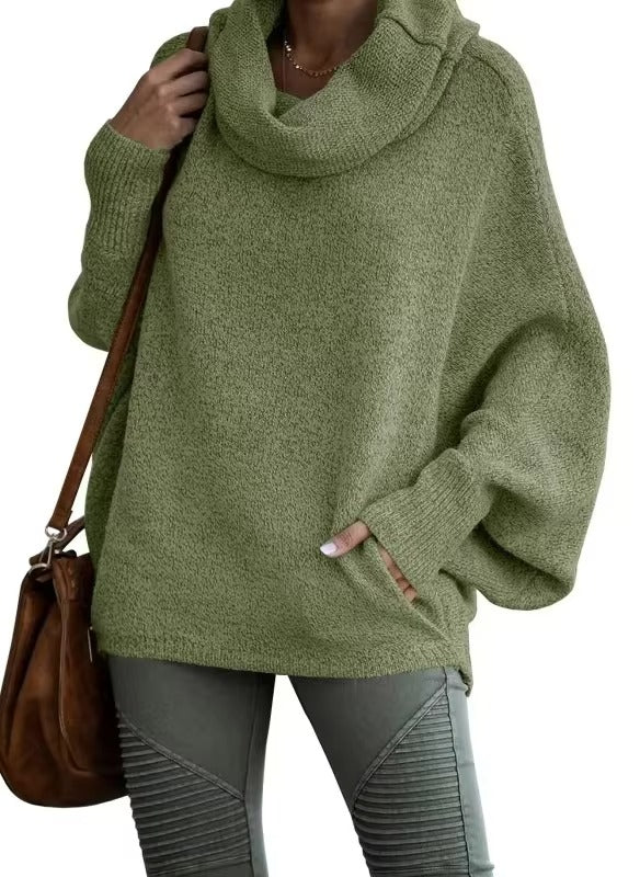 Une femme porte un pull vert sauge à manches chauve-souris oversize et col vert. La matière douce et confortable crée une sensation de cocon douillet. Elle associe ce vêtement à un pantalon kaki texturé et un sac en cuir marron, pour un look hivernal à la fois naturel et élégant.
