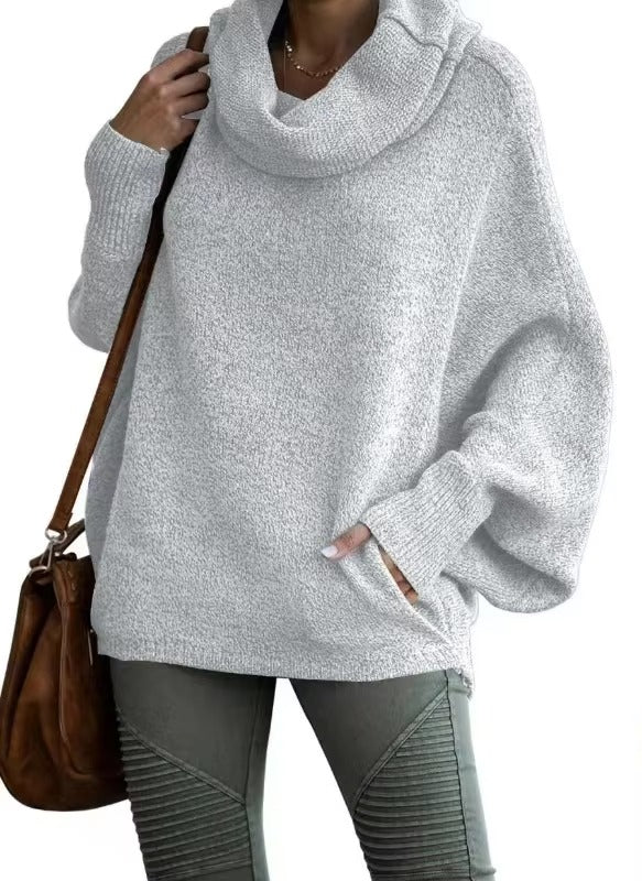 Femme vêtue d’un pull ample à manches chauve-souris de couleur gris clair avec un large col drapé. Le tricot souple et doux apporte une allure cocooning et élégante. Elle l’associe à un pantalon kaki texturé et un sac en cuir marron, parfait pour un look d’hiver décontracté et moderne.