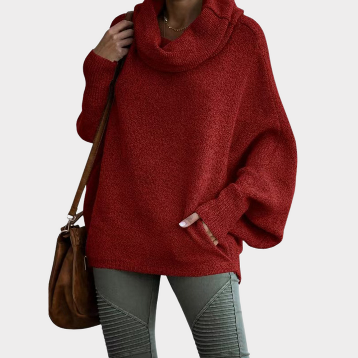 Une femme porte un pull rouge oversize à manches chauve-souris et à large col drapé. La maille douce et épaisse lui assure confort et chaleur pour les mois les plus froids. Elle complète sa tenue avec un pantalon kaki texturé et un sac en cuir marron pour un look hivernal à la fois élégant et décontracté.