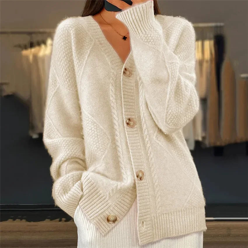 Femme portant un cardigan en tricot beige à col en V, avec des boutons en bois et un design torsadé. Elle tient une main près du visage tout en affichant un look élégant.