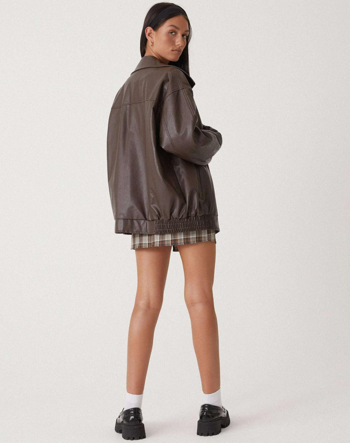 Vue arrière de la veste oversize en PU pour femme, modèle Ombeline. Veste en simili-cuir marron à la coupe ample et taille élastiquée, portée avec une jupe à carreaux et des chaussures noires. Un look moderne et confortable, parfait pour l'hiver.