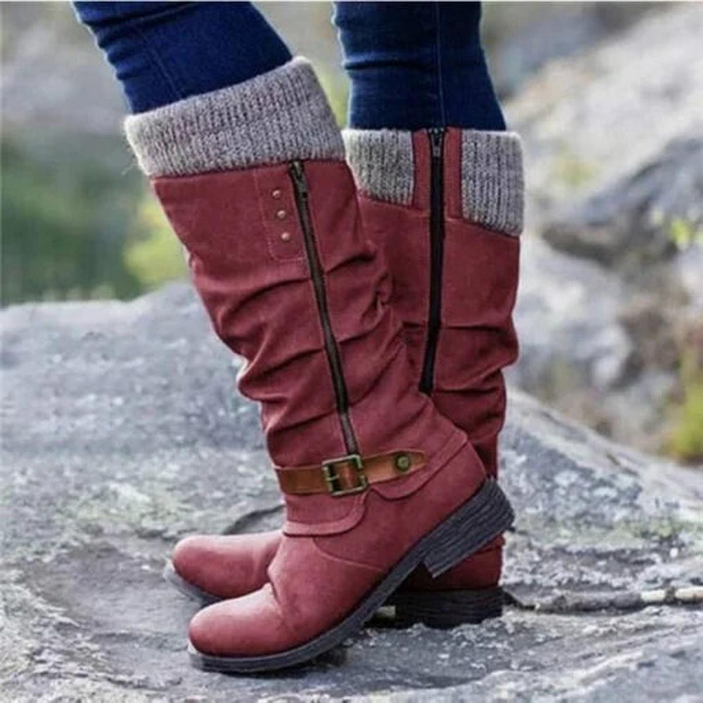 Bottes hautes pour femme de couleur rouge bordeaux, avec revers tricoté beige en haut et fermeture éclair latérale. Le design présente un effet plissé raffiné et une sangle marron à boucle métallique autour de la cheville pour une touche élégante. Portées avec un jean ajusté, ces bottes au style moderne et confortable offrent une semelle épaisse antidérapante, idéales pour la saison automne-hiver. La photo est prise en extérieur sur un terrain rocheux, soulignant leur aspect pratique et tendance.