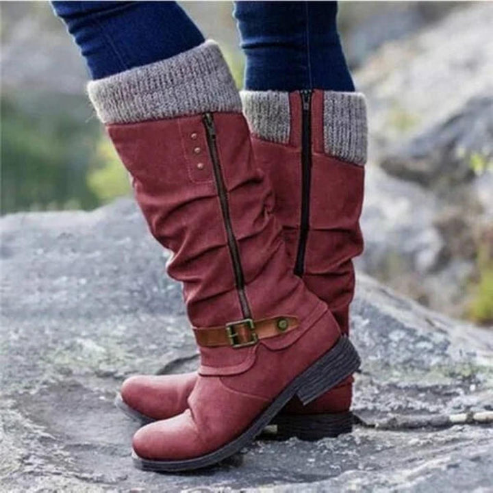 Bottes hautes pour femme de couleur rouge bordeaux, avec revers tricoté beige en haut et fermeture éclair latérale. Le design présente un effet plissé raffiné et une sangle marron à boucle métallique autour de la cheville pour une touche élégante. Portées avec un jean ajusté, ces bottes au style moderne et confortable offrent une semelle épaisse antidérapante, idéales pour la saison automne-hiver. La photo est prise en extérieur sur un terrain rocheux, soulignant leur aspect pratique et tendance.