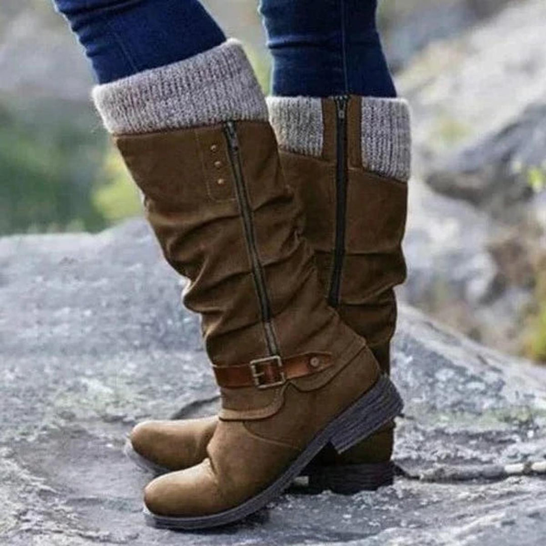 Bottes hautes pour femme de couleur marron, dotées d’un revers tricoté beige sur le haut et d’une fermeture éclair latérale. Le design présente un effet plissé élégant et une sangle décorative en cuir marron avec boucle métallique au niveau de la cheville. Portées avec un jean ajusté, ces bottes au style moderne et confortable disposent d’une semelle épaisse antidérapante, idéales pour les saisons fraîches. La photo, prise en extérieur sur un sol rocheux, met en valeur leur allure tendance et pratique.