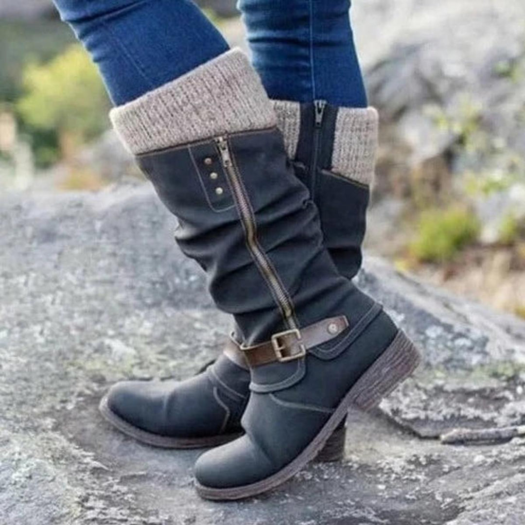 Bottes hautes pour femme de couleur noire, avec un revers tricoté beige en haut et une fermeture éclair latérale. Le design présente un effet plissé moderne et une sangle marron à boucle métallique au niveau de la cheville, ajoutant une touche élégante et décontractée. Portées avec un jean ajusté, ces bottes au style pratique offrent une semelle épaisse antidérapante, parfaites pour affronter la saison froide avec confort et style. La photo est prise en extérieur sur un sol rocheux.