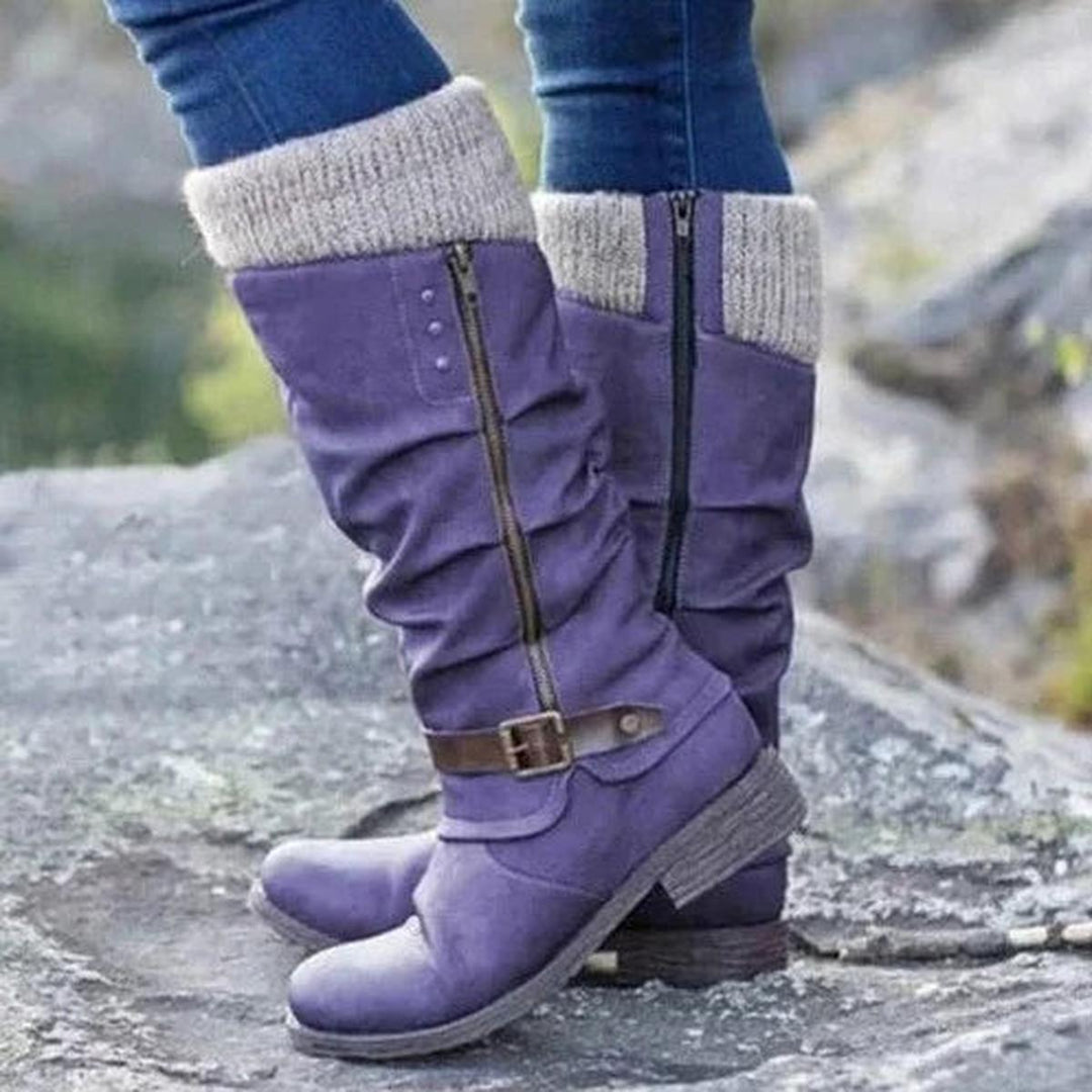Bottes hautes pour femme de couleur violette, ornées d’un revers tricoté beige sur le haut et d’une fermeture éclair latérale. Le design présente un effet plissé élégant et une sangle marron avec boucle métallique autour de la cheville pour une touche chic. Portées avec un jean ajusté, ces bottes au style moderne et confortable sont dotées d’une semelle épaisse antidérapante, parfaites pour les journées fraîches. La photo est prise en extérieur sur un sol rocheux, mettant en valeur leur look tendance et pra