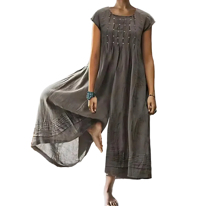 Femme portant une combinaison une-pièce fluide de couleur gris taupe, à manches courtes et coupe ample, avec un tissu léger et aérien qui tombe jusqu’aux chevilles, présentée dans un intérieur lumineux au style minimaliste.