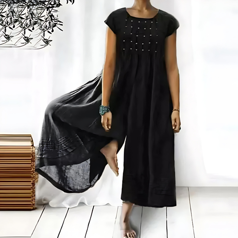 Femme portant une combinaison une-pièce noire fluide, à manches courtes et coupe ample, avec de délicats détails décoratifs sur le buste, confectionnée dans un tissu léger et aérien tombant jusqu’aux chevilles, présentée dans un intérieur lumineux et épuré.